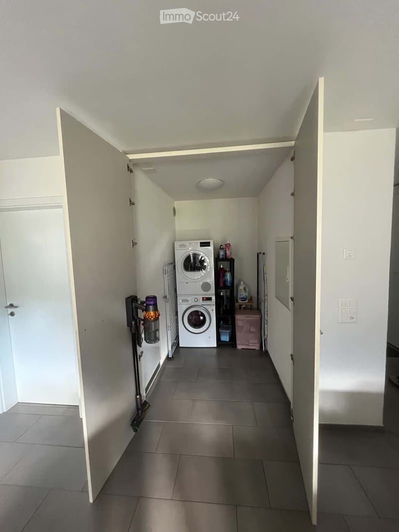 A narrow hallway with tiled flooring and white walls, leading to a laundry area with a washing machine, dryer, and cleaning supplies.