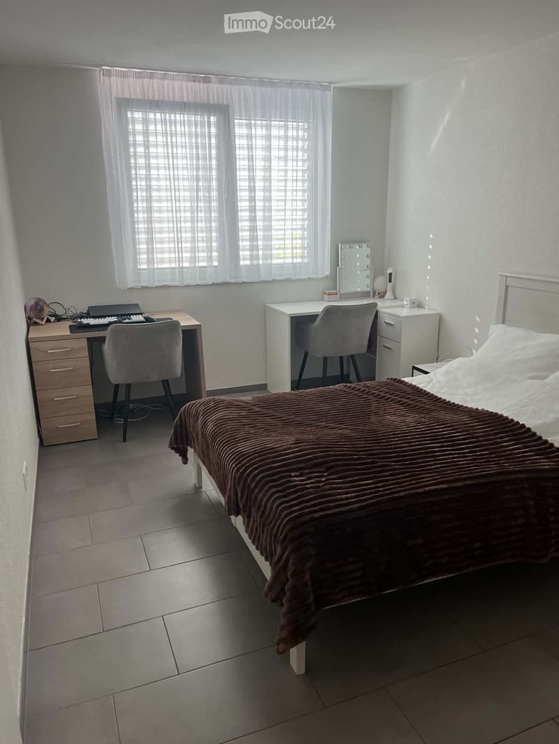 A modern bedroom with a large bed, a desk with a laptop and keyboard, a vanity with a mirror, and two chairs.