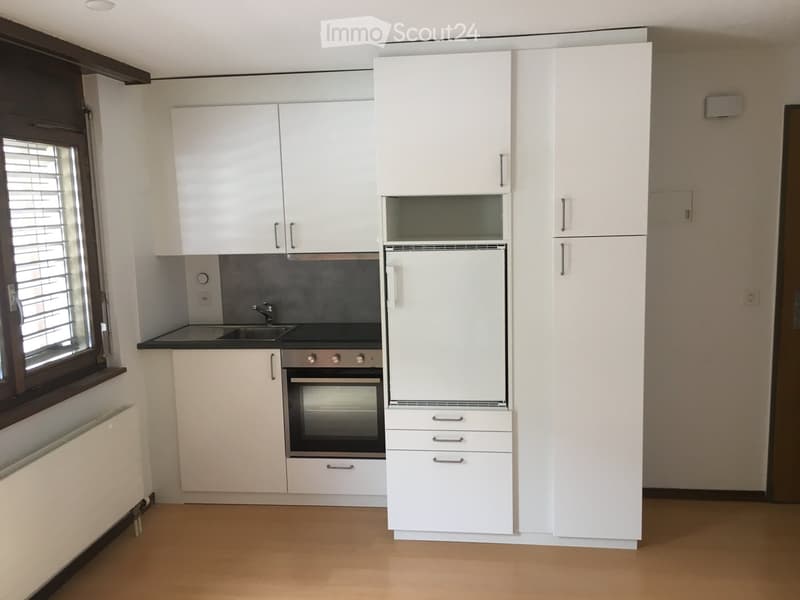 Modern kitchen with white cabinets, stainless steel oven, refrigerator, and sink, featuring a window with blinds and a radiator below.