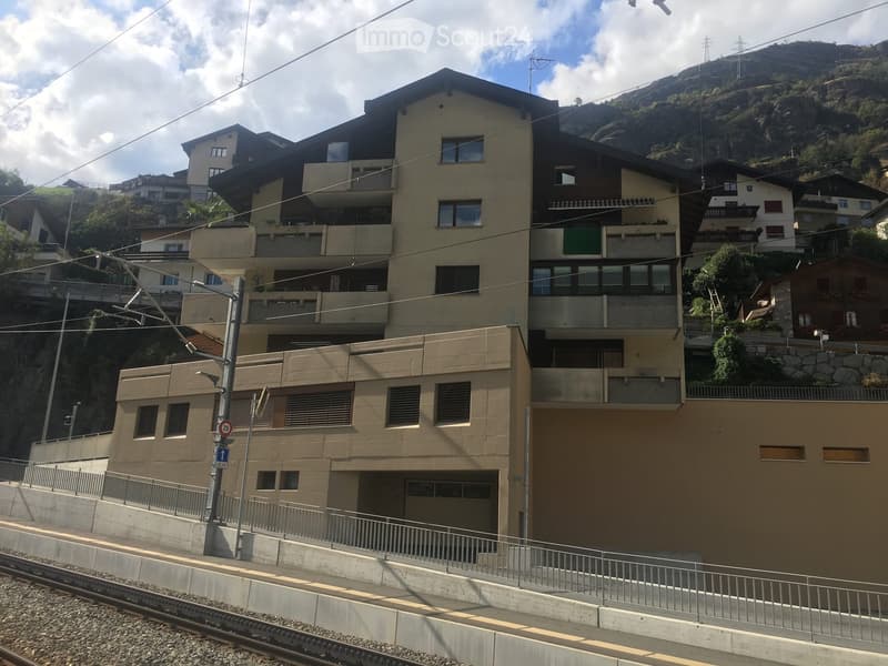 Multi-story residential building with balconies and large windows, situated near a railway track with a mountainous backdrop.