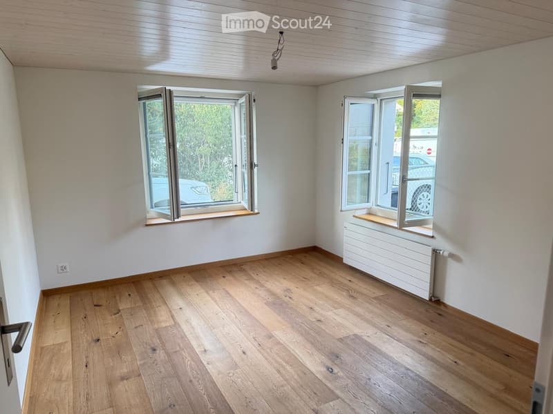 Empty room with light wooden flooring, white walls, and large windows with wooden sills. Radiator visible under one window.