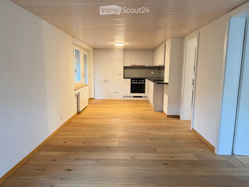 Empty hallway with wooden flooring, white walls, and a small kitchen area visible at the end. The kitchen features white cabinets, a stainless steel oven, and a countertop.