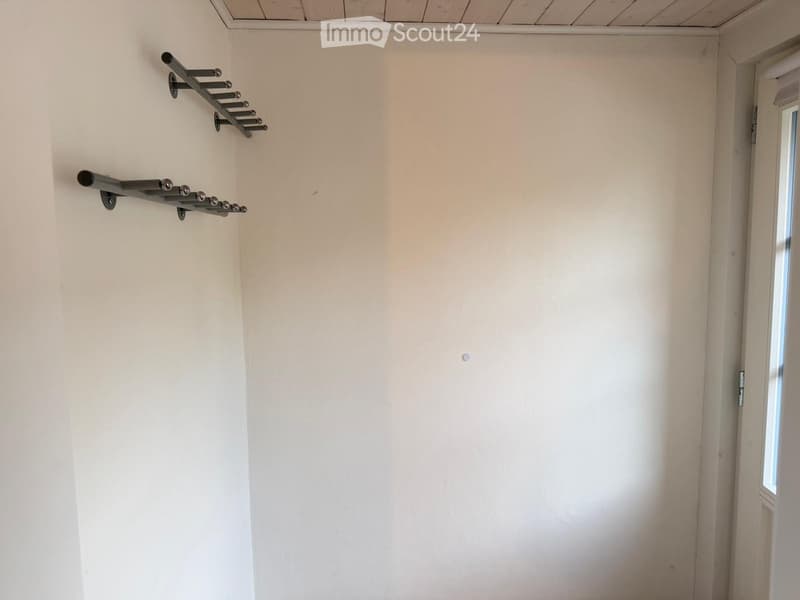 Empty room with white walls, wooden ceiling, and a small window. Metal coat rack with hooks is mounted on the wall.