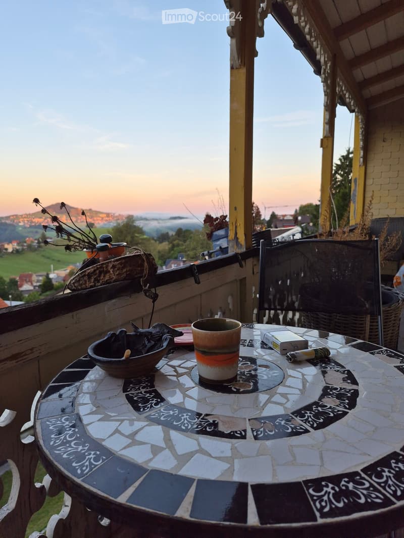 Balcony with a mosaic table, a cup, a bowl, a book, and a view of the countryside at sunset.