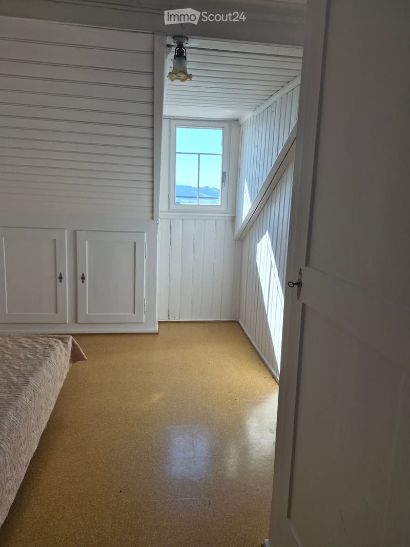 A narrow hallway with a bed on the left side, white paneled walls, and a window at the end. The floor is a light-colored, possibly laminate material.