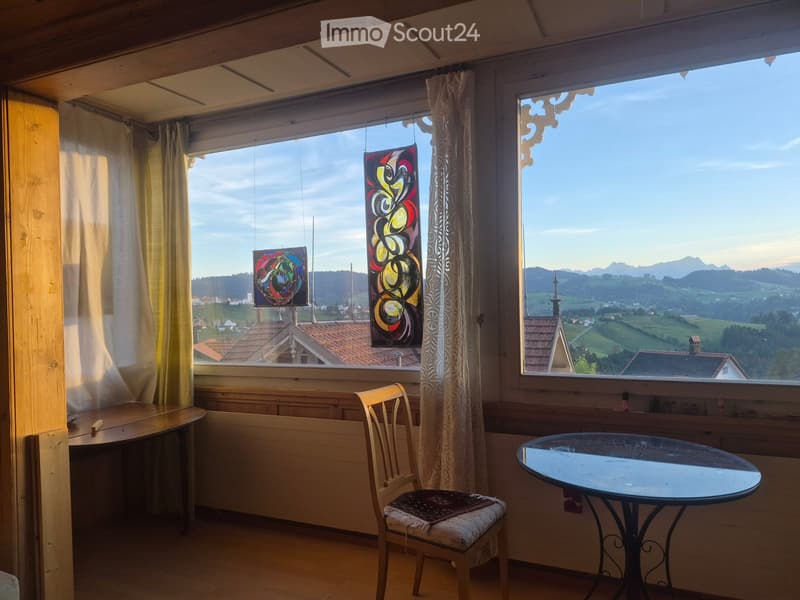 Bright room with large windows offering a scenic mountain view, featuring a wooden chair, a round glass table, and decorative curtains.