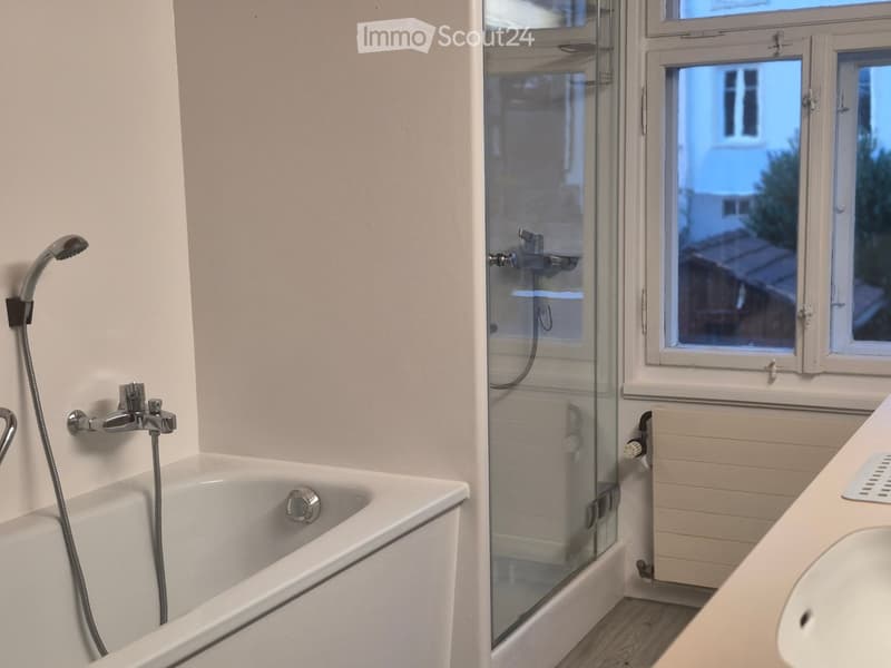 Modern bathroom with a bathtub, glass shower enclosure, sink, and radiator. Features a chrome faucet and showerhead, with large windows providing natural light.