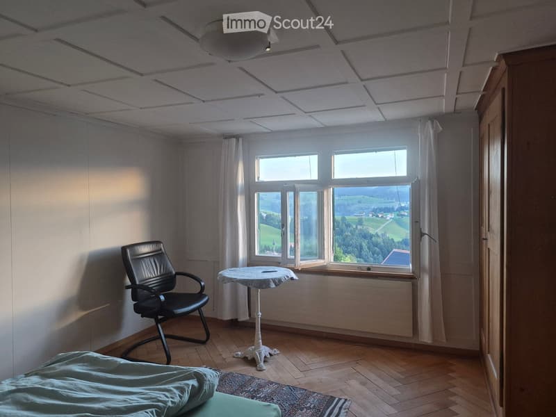 Bright room with wooden flooring, large windows offering scenic views, a bed with a blue blanket, a black chair, and a small round table.