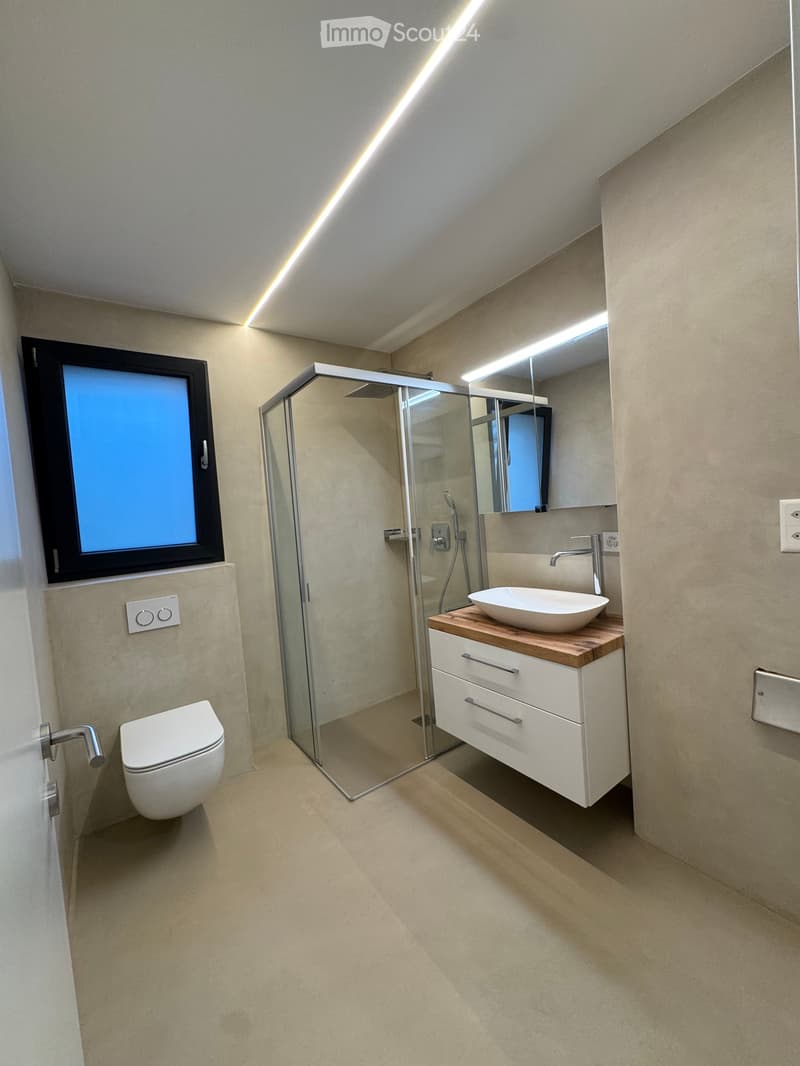 Modern bathroom with a toilet, sink, mirror, and glass-enclosed shower stall. Features a sleek design with light-colored tiles and a wooden vanity.