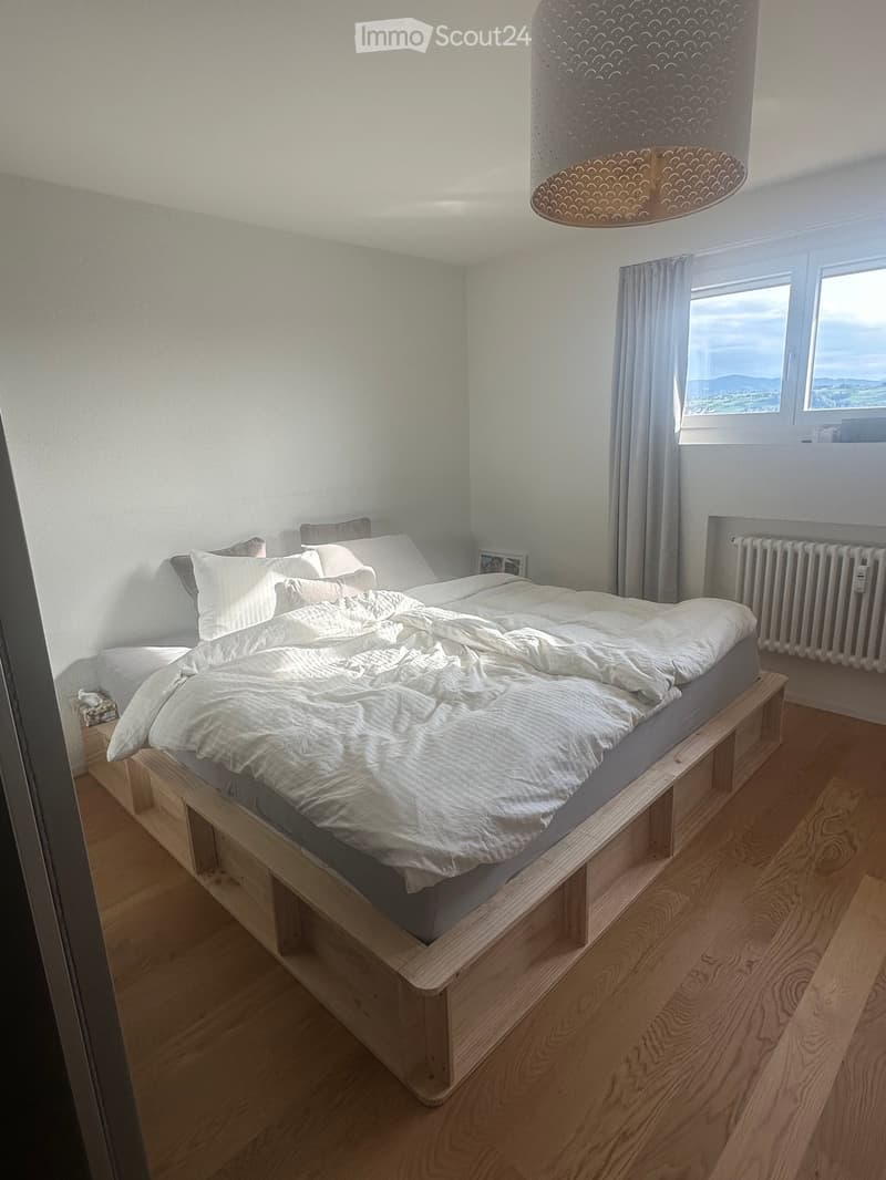 A modern bedroom with a wooden bed frame, white bedding, a large window with curtains, a radiator, and a decorative hanging lamp.