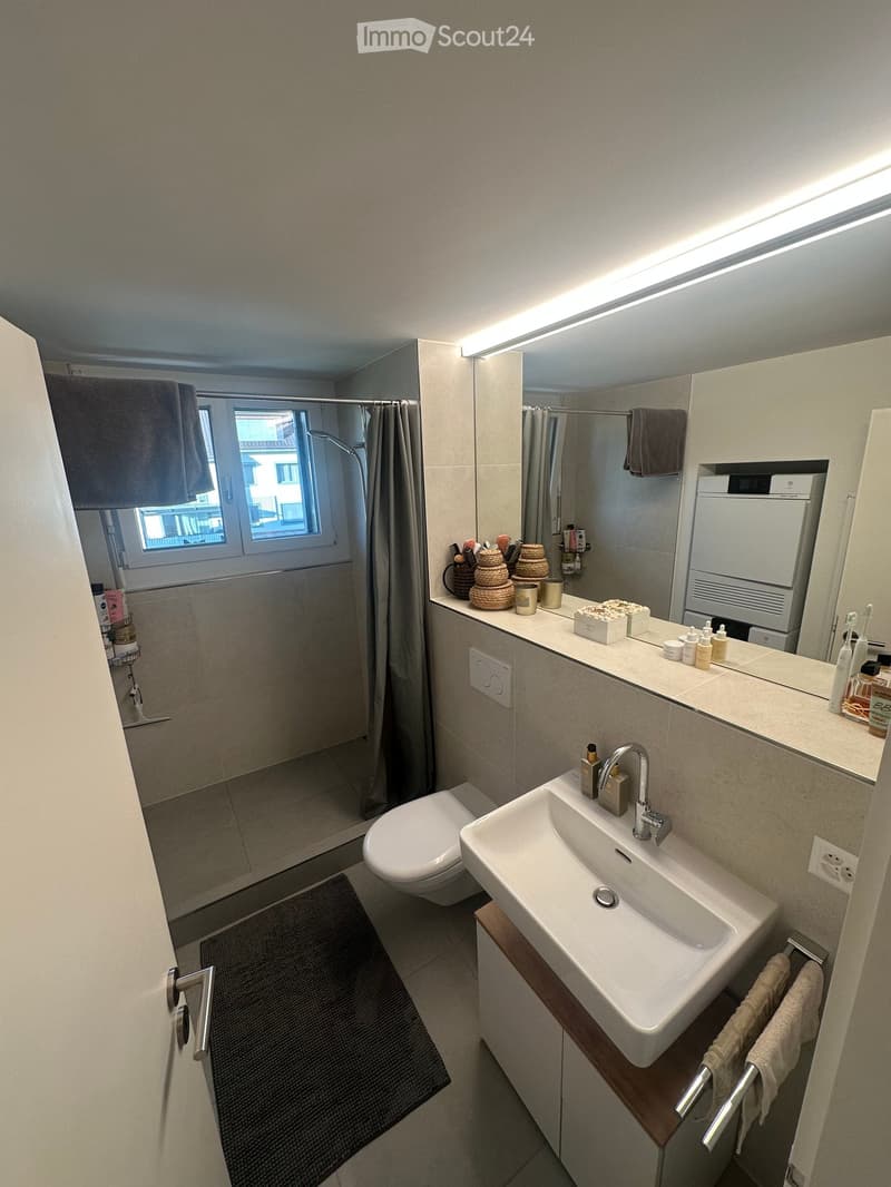 Modern bathroom with a white sink, chrome faucet, toilet, large mirror, and a shower area with a window and curtain. The countertop holds various toiletries and decorative items.