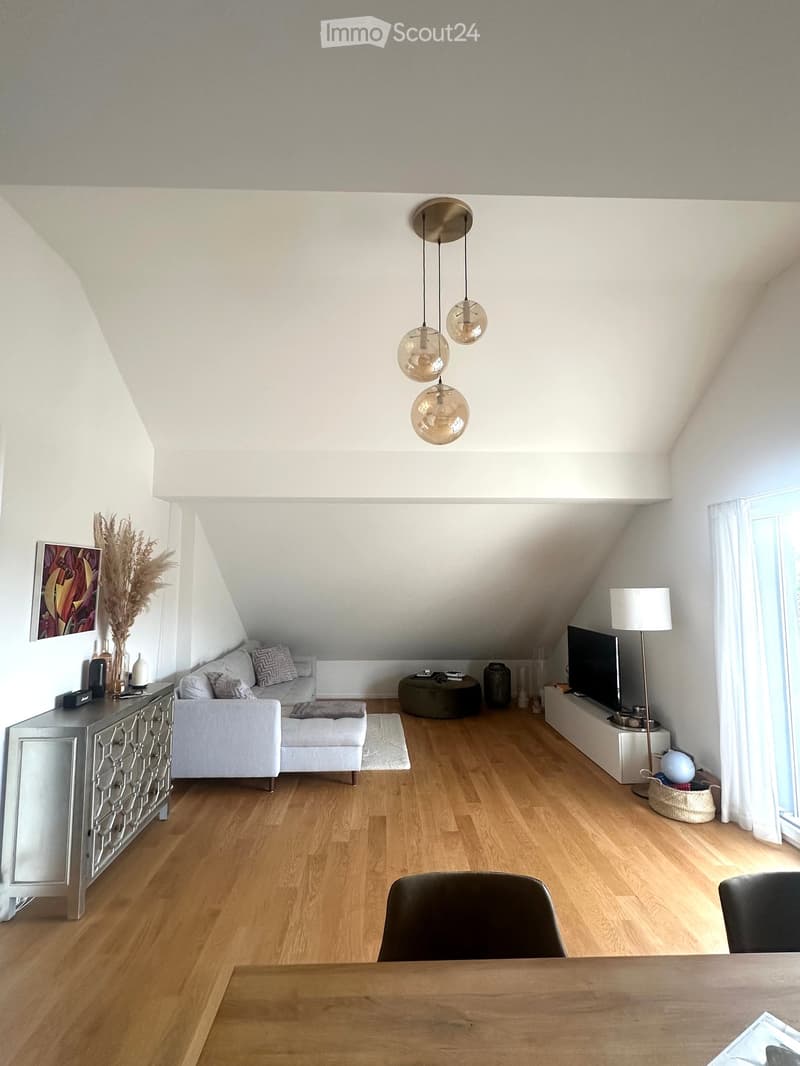 Bright living room with wooden flooring, white walls, and a slanted ceiling. Features a grey sofa with an ottoman, a wooden dining table with chairs, a silver cabinet, a floor lamp, and a pendant light fixture.