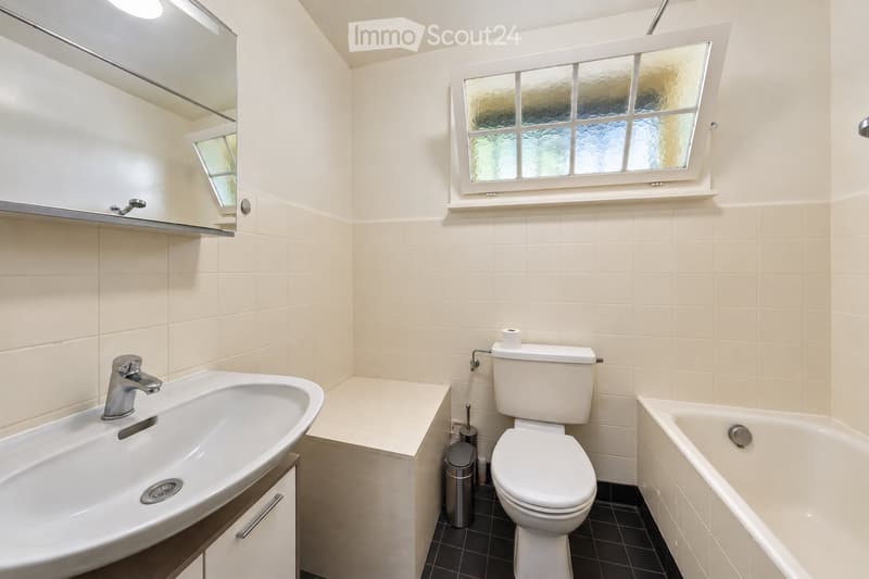 Modern bathroom with white tiles, a sink, toilet, bathtub, and a mirror above the sink. Features a window with frosted glass and a small shelf above the sink.