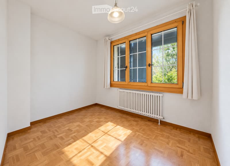 Empty room with wooden parquet flooring, white walls, large wooden-framed windows with white curtains, and a white radiator below the windows.