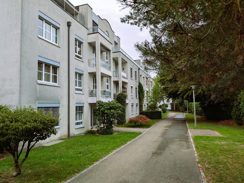 Modern multi-story residential building with balconies, surrounded by greenery and a paved pathway.