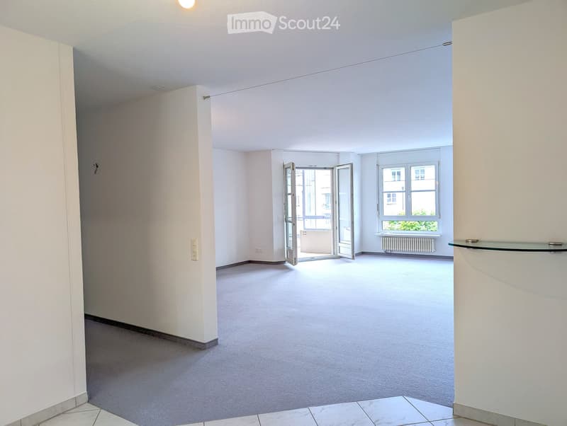 Empty room with light-colored walls, carpeted floor, and a large window with a radiator below. An open door leads to a balcony area.