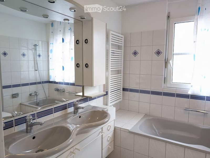 Modern bathroom with two sinks, a bathtub, a large mirror, a radiator, and a window with a curtain.