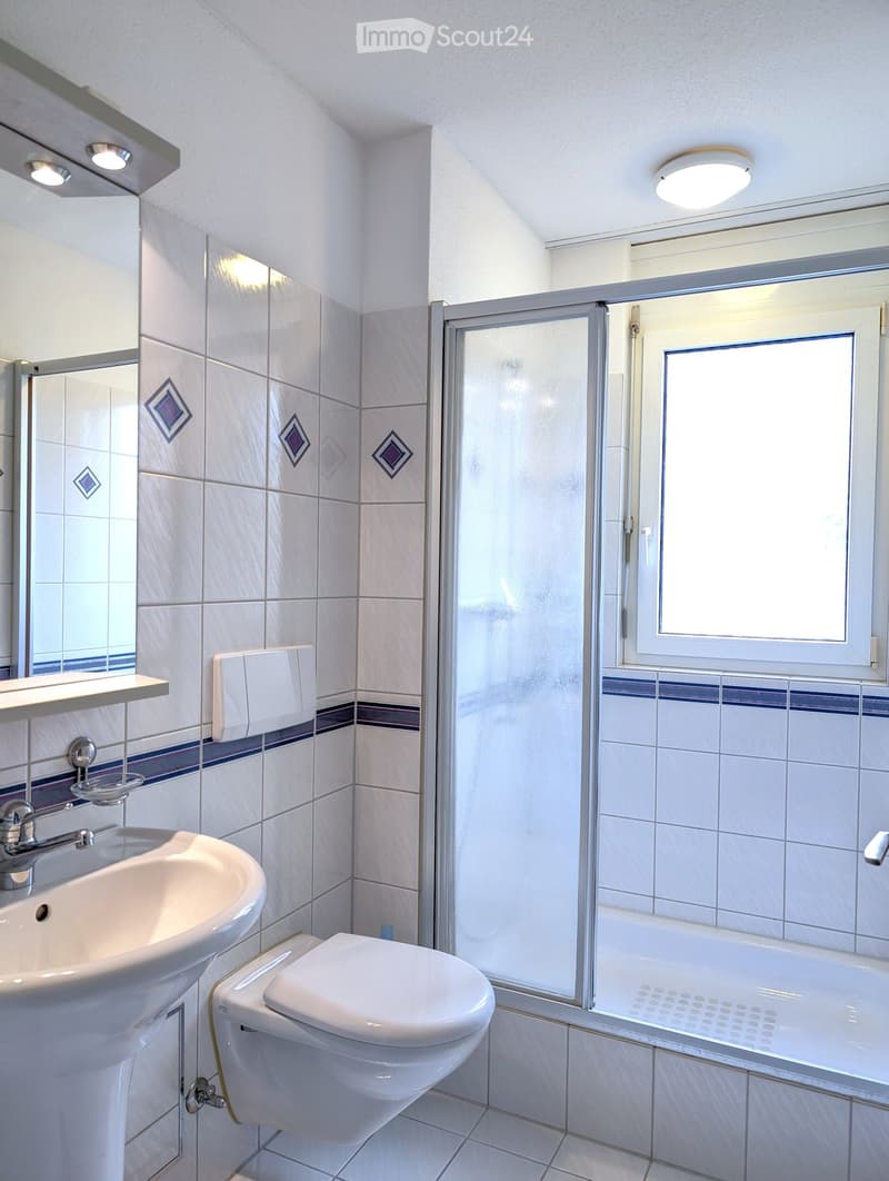 Modern bathroom with white tiles, a sink, toilet, bathtub, and a glass-enclosed shower. Features a window and decorative tiles on the walls.