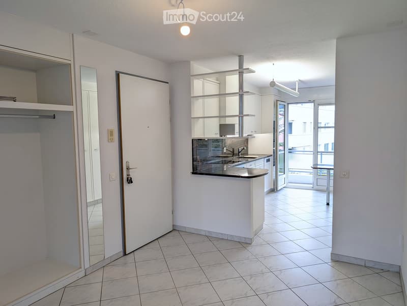 Modern, minimalist kitchen with white cabinets, a central island, and a sink. A mirror and a small table are visible, with a door leading to a balcony.