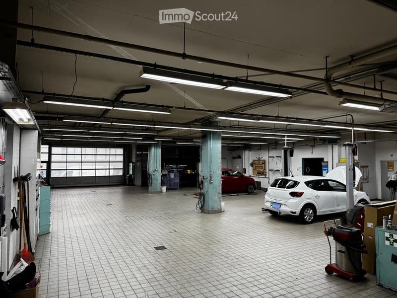 A spacious garage with tiled flooring, fluorescent lighting, and two cars (one red and one white) positioned on hydraulic lifts. Cleaning equipment and tools are visible along the walls.