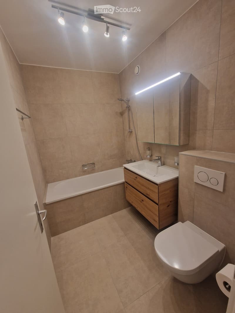 Modern bathroom with beige tiles, a bathtub, a sink with wooden cabinet, a toilet, and a large mirror with built-in lighting.