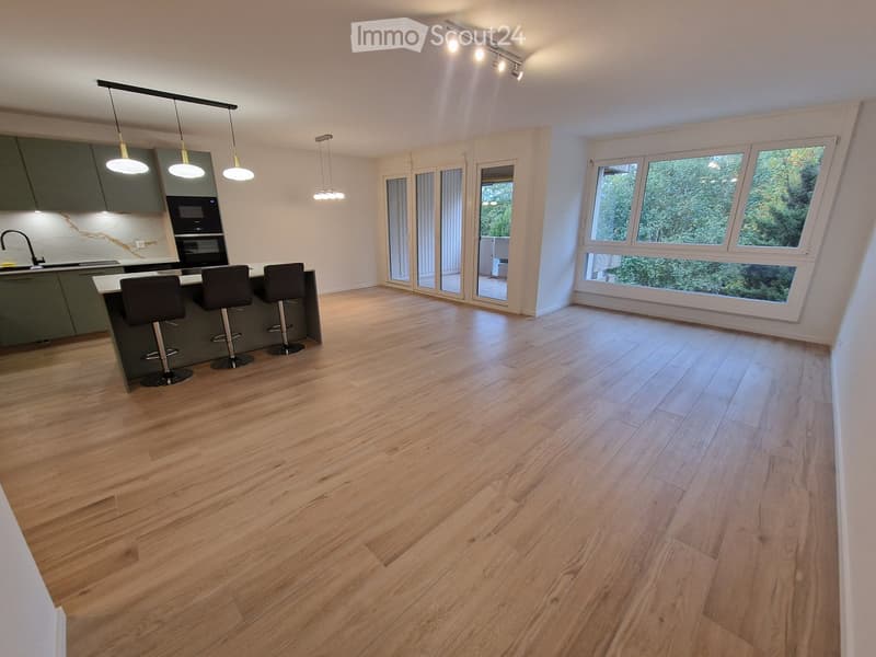 Modern kitchen with light wooden flooring, grey cabinets, island with bar stools, and large windows overlooking greenery.