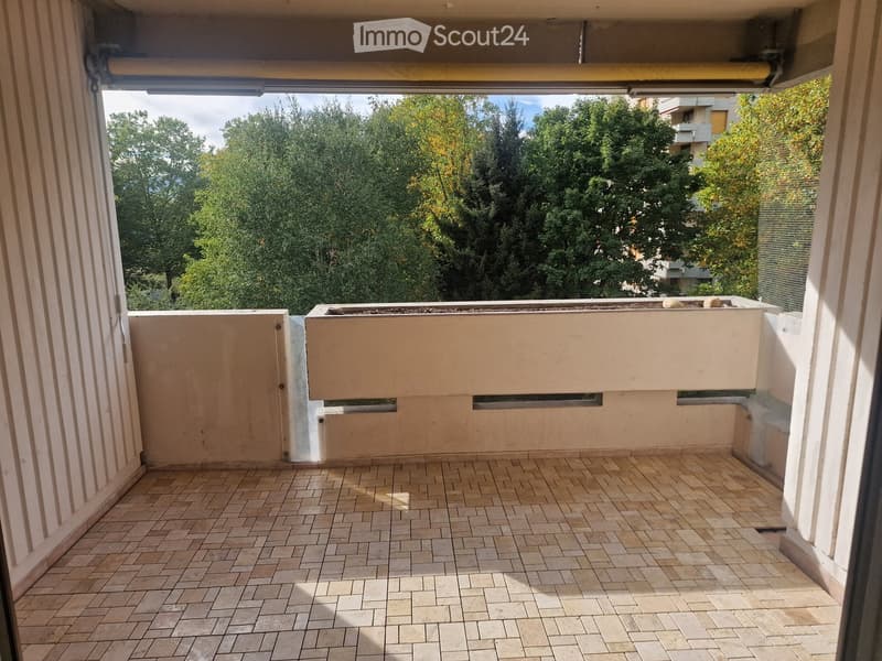 Balcony with tiled floor, low wall, and small planter box, overlooking a green area with trees and buildings.