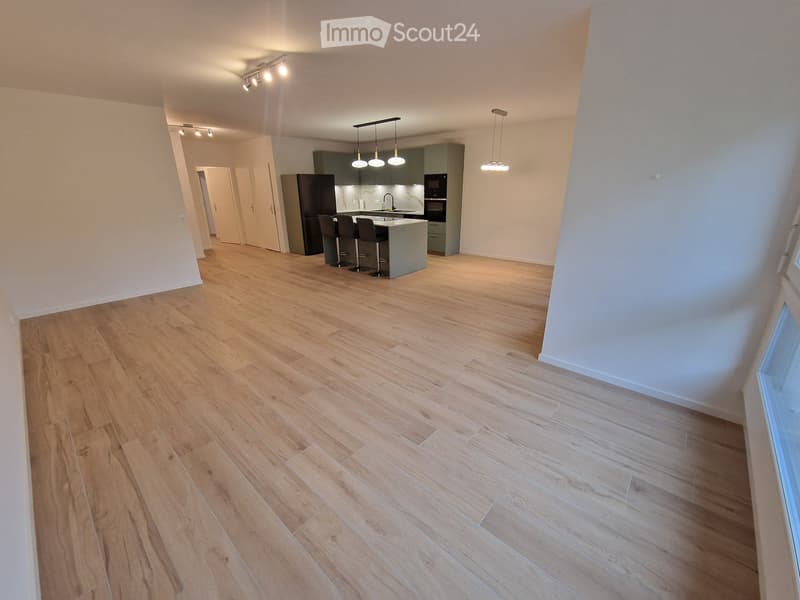 Empty room with wooden flooring, white walls, and a small kitchen area in the background.