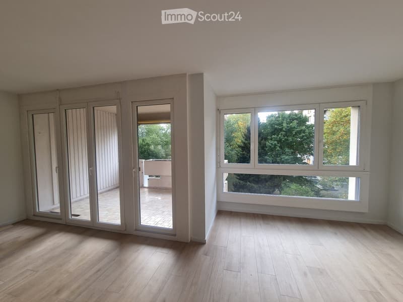 Empty room with wooden flooring, large windows, and sliding glass doors leading to a balcony.