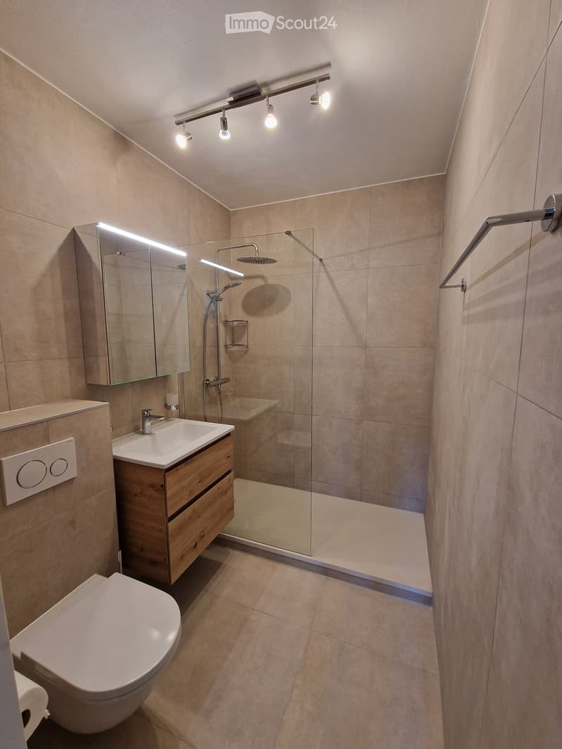 Modern bathroom with beige tiles, a toilet, a sink with a wooden cabinet, a glass-enclosed shower, and a mirror with built-in lighting.