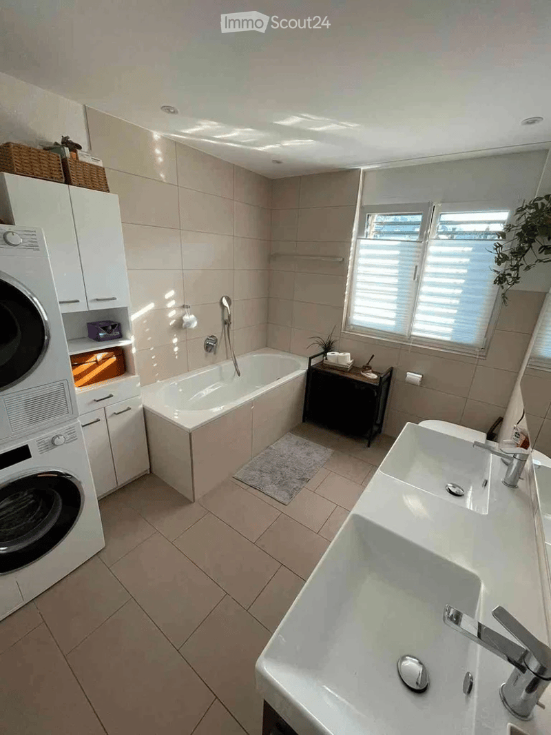 Modern bathroom with a bathtub, double sink, washing machine, and a small plant on a side table. The space features light-colored tiles and a window with blinds.
