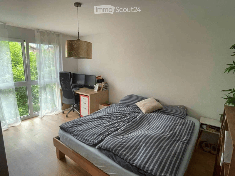 A well-lit bedroom with a double bed, a desk with a computer, and a hanging lamp. The room features large windows with sheer curtains and a wooden floor.