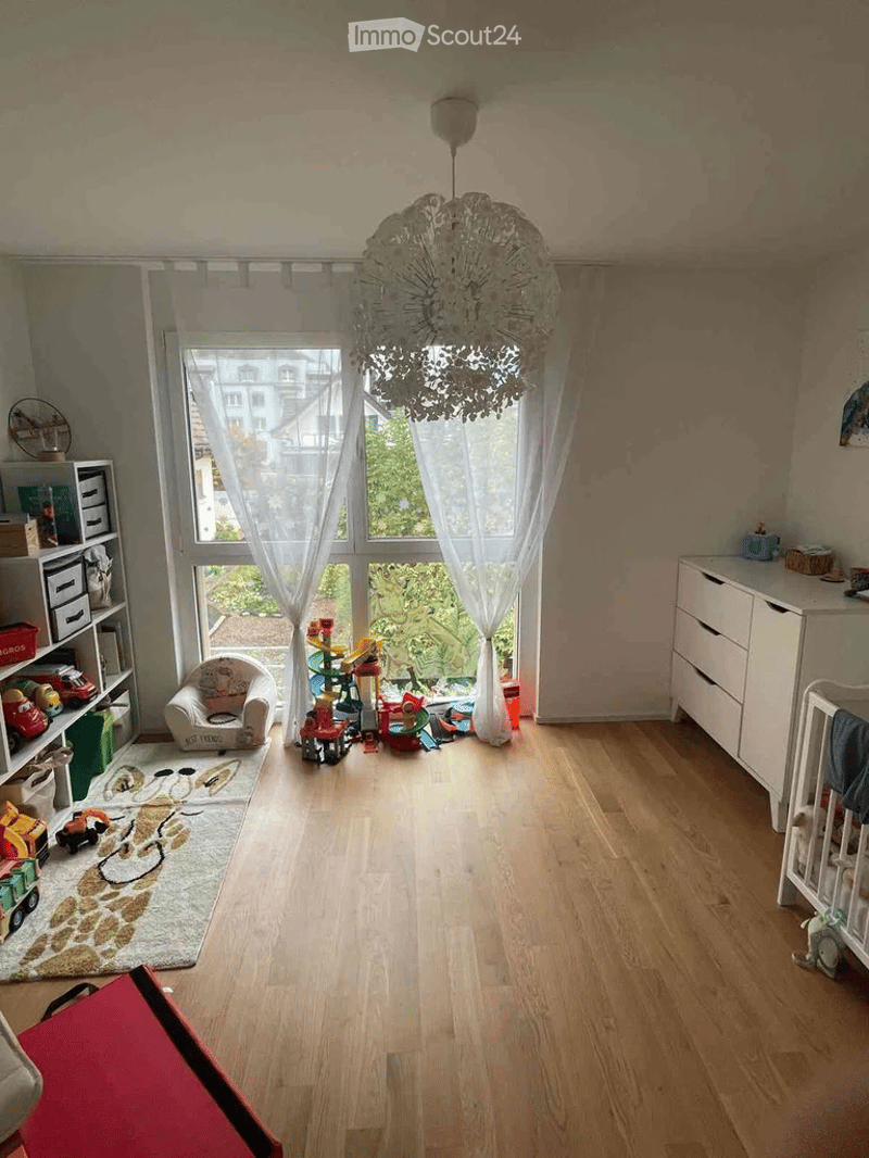 Brightly lit children's room with wooden flooring, large window with sheer curtains, a small armchair, shelves filled with toys, a crib, and a colorful rug with a bunny design.