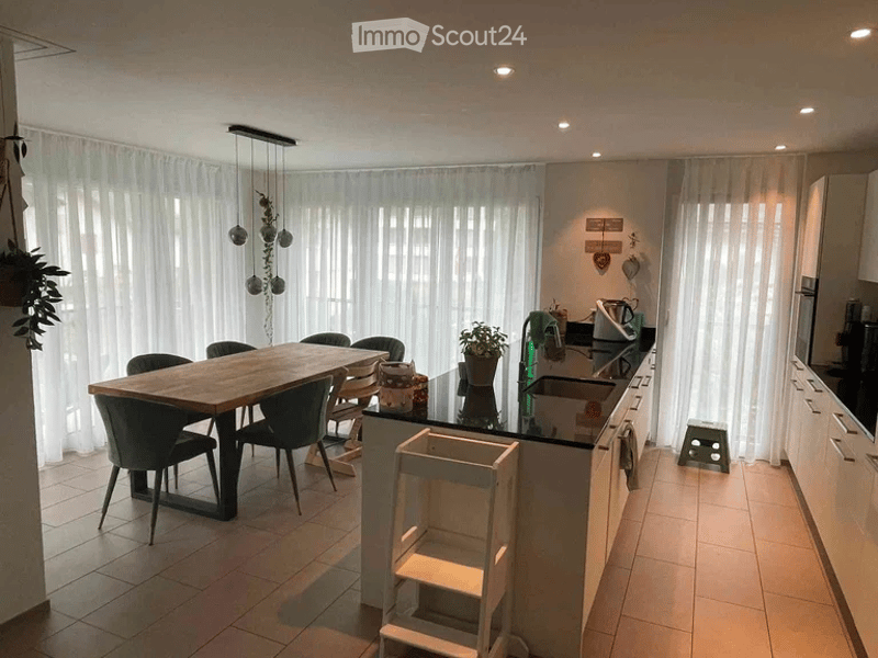 Modern kitchen with a wooden dining table and chairs, a kitchen island with a sink, and white cabinets. The space features large windows with sheer curtains and pendant lights.