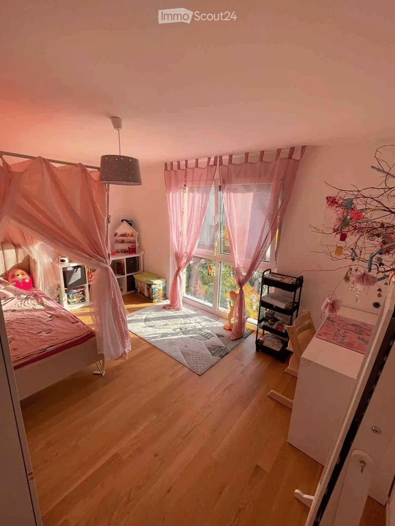 Brightly lit bedroom with wooden flooring, large window with pink curtains, bed with pink bedding, hanging canopy, small table, and various toys and storage items.