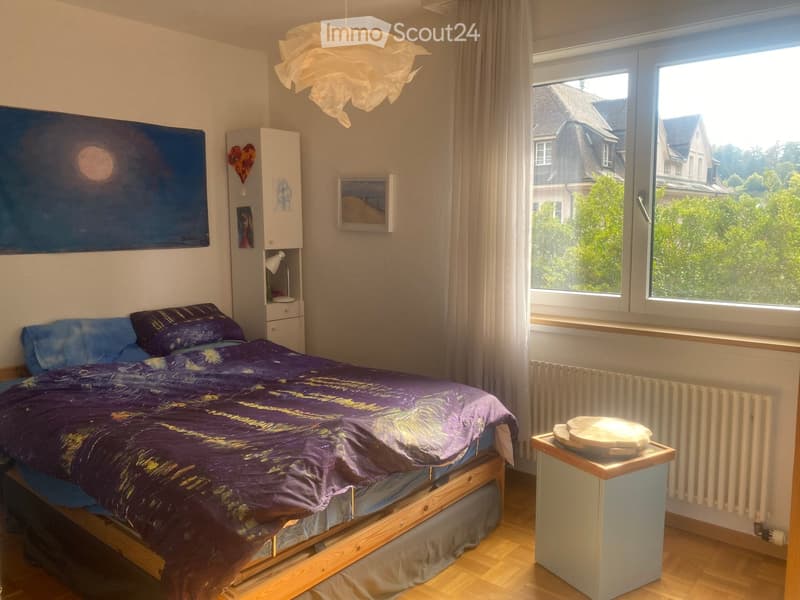 Cozy bedroom with a wooden bed, purple bedding, nightstand, lamp, and a large window with a view of the neighborhood.