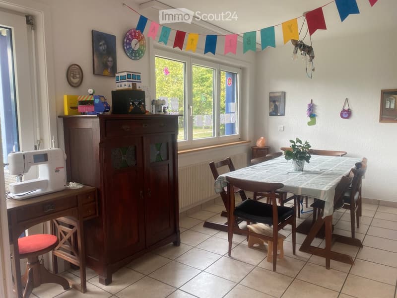 Bright room with a dining table and chairs, a wooden cabinet, a sewing machine on a desk, and various decorative items including a clock and artwork.