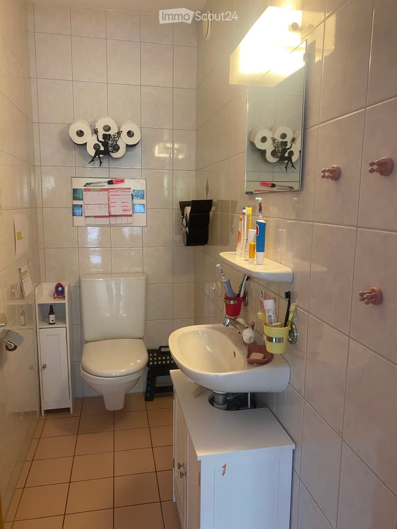 A small bathroom with a toilet, sink, mirror, and various toiletries. The walls are tiled, and there is a small cabinet and a toothbrush holder visible.