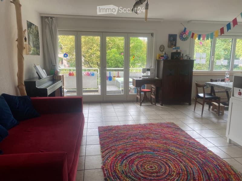 Bright living room with large sliding glass doors leading to a balcony, a red sofa, a colorful circular rug, a piano, a small table with chairs, and a cabinet.