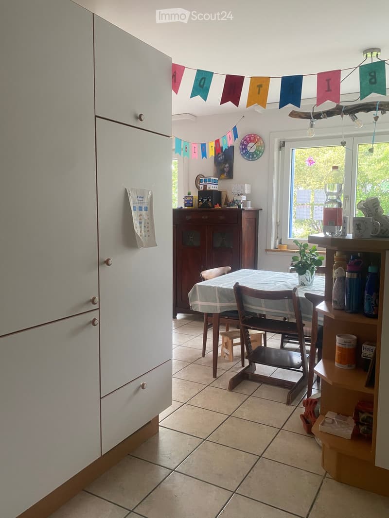 Bright kitchen with tiled floor, white refrigerator, wooden cabinet, dining table with chairs, and various kitchen items on shelves and counters.