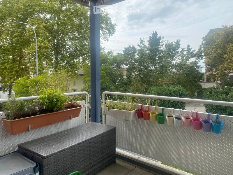 Balcony with a wicker storage box, hanging planters, and colorful hanging pots, surrounded by trees and greenery.