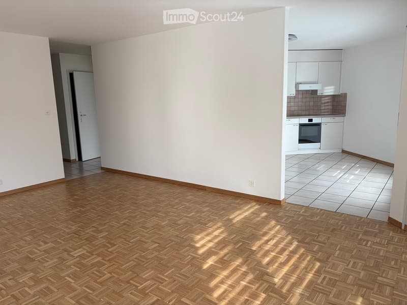 Empty room with wooden herringbone flooring and white walls, connected to a tiled kitchen area with white cabinets and appliances.
