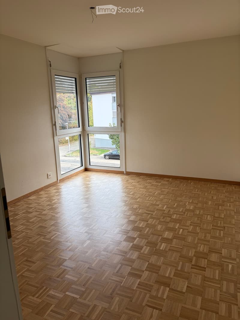 Empty room with wooden herringbone flooring, large windows with blinds, and light-colored walls.