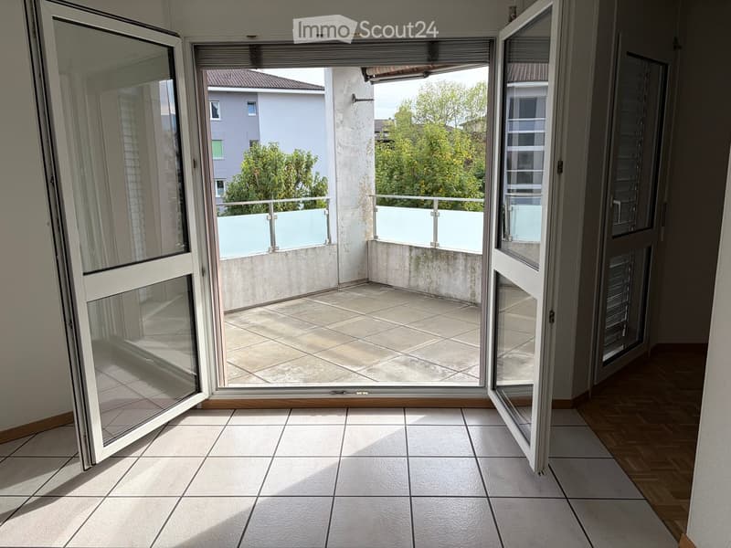 Open sliding doors lead to a tiled balcony with a low white railing and a view of neighboring buildings and greenery.