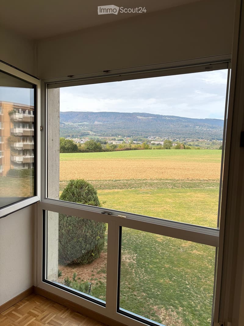 Empty room with wooden flooring and large windows offering a scenic view of a green field, distant hills, and a neighboring building.