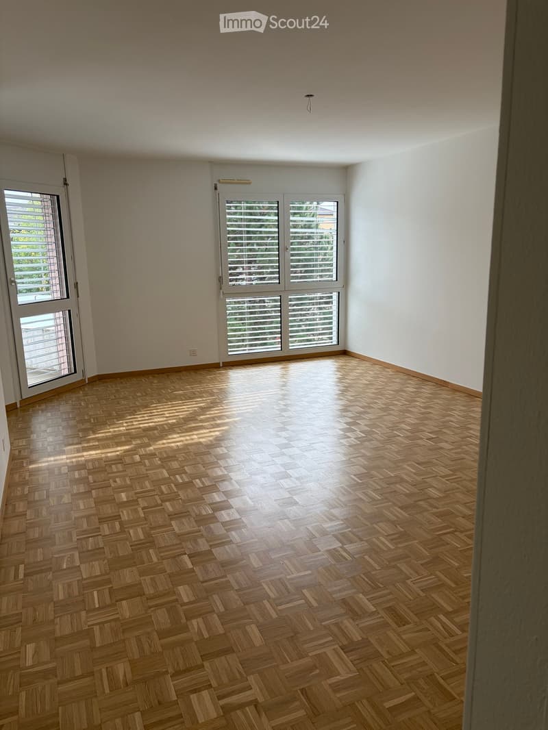 Empty room with wooden parquet flooring, white walls, and large windows with blinds. No furniture or equipment present.