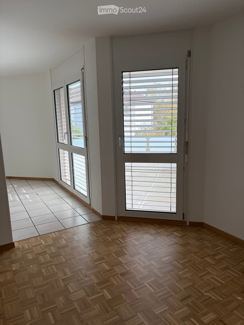 Empty room with wooden herringbone flooring, large windows with blinds, and a sliding glass door leading to a tiled area.