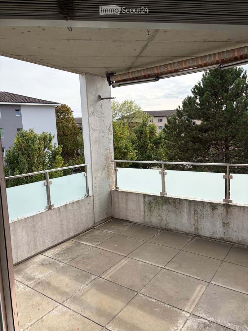 Balcony with tiled floor, glass railing, and covered roof, overlooking a residential area with trees and houses.