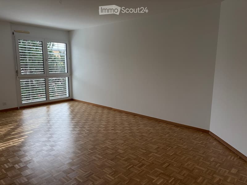 Empty room with wooden herringbone flooring, white walls, and large windows with blinds.