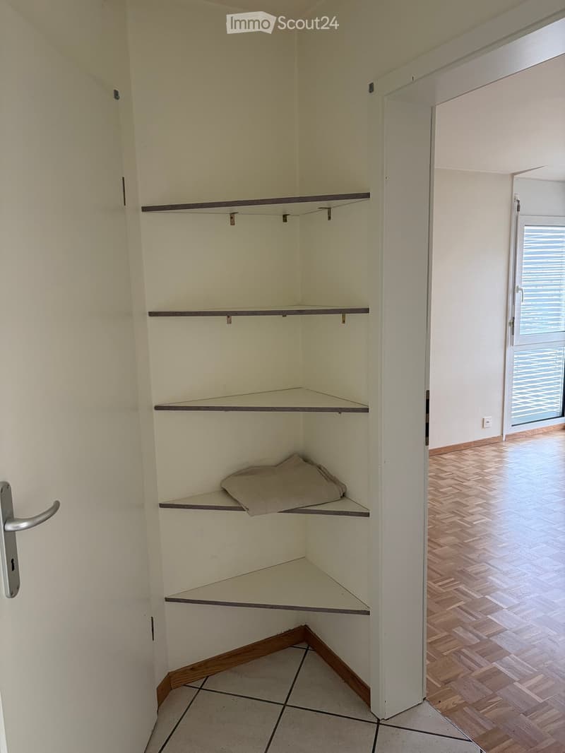 Empty corner closet with built-in shelves and a single folded blanket on one shelf. The room has tiled flooring and connects to another area with wooden flooring and a window with blinds.